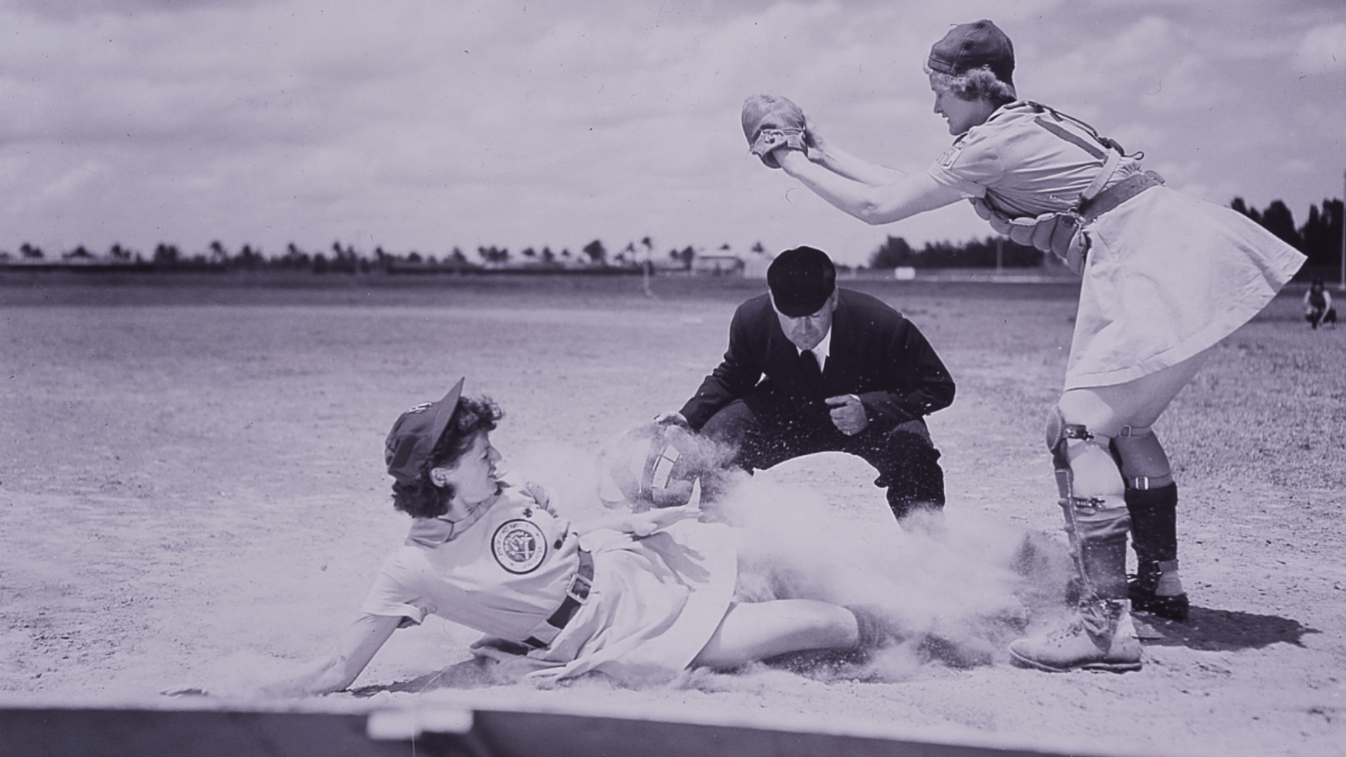 Two women playing baseball. One sliding into a base while the other catches the ball. An umpire stands behind them to make a call on the play.
