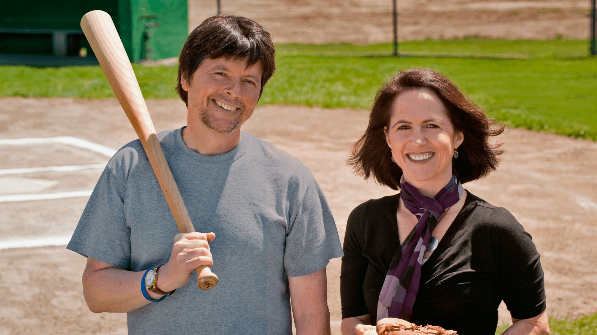 A man (left) and woman (right) standing outdoors. The man is standing casually with a wooden baseball bat.
