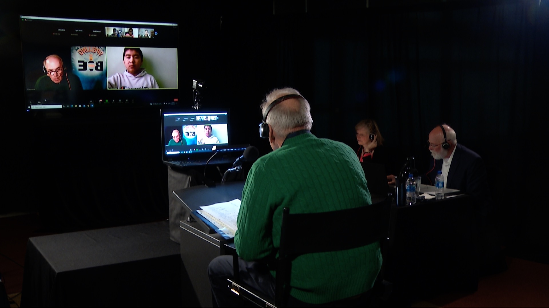 A man in a green sweater sits in front of a laptop during a television production. His back is facing the camera. He is speaking with a young man via Zoom.