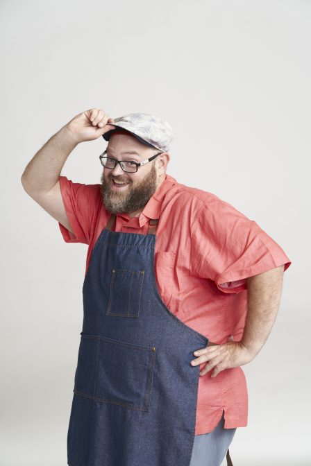 A man in an apron stands in front of a white background with a hand on his hip and his ball cap.