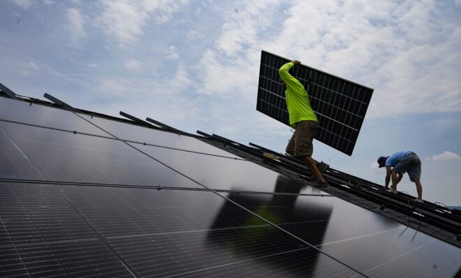 FILE - Nicholas Hartnett, owner of Pure Power Solar, carries a panel as he and Brian Hoeppner, right, install a solar array on the roof of a home in Frankfort, Ky., Monday, July 17, 2023. President Joe Biden has been careful not to declare an outright victory against inflation, but the White House says the cost savings from the Inflation Reduction Act are coming as the law is getting enacted. Tax credits will reduce the cost of installing rooftop solar panels by 30%, which will in turn lower monthly electricity bills. (AP Photo/Michael Conroy, File)